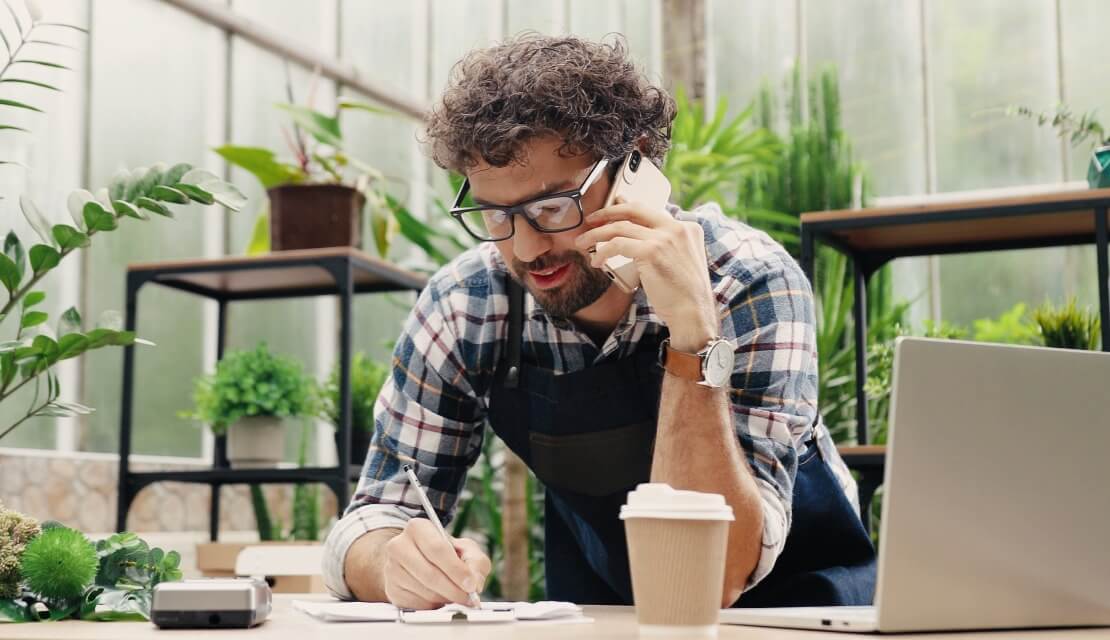 Plant shop owner at his work desk talking on phone and taking down an order.