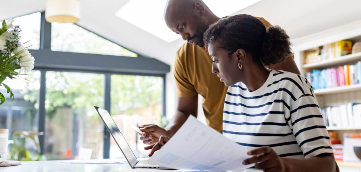 Woman holding paper and looking at her computer next to a man, holding a credit card.
