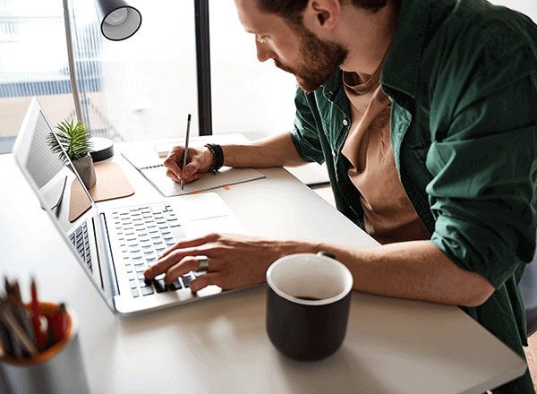 Man sitting in front of a laptop and writing notes on a note pad