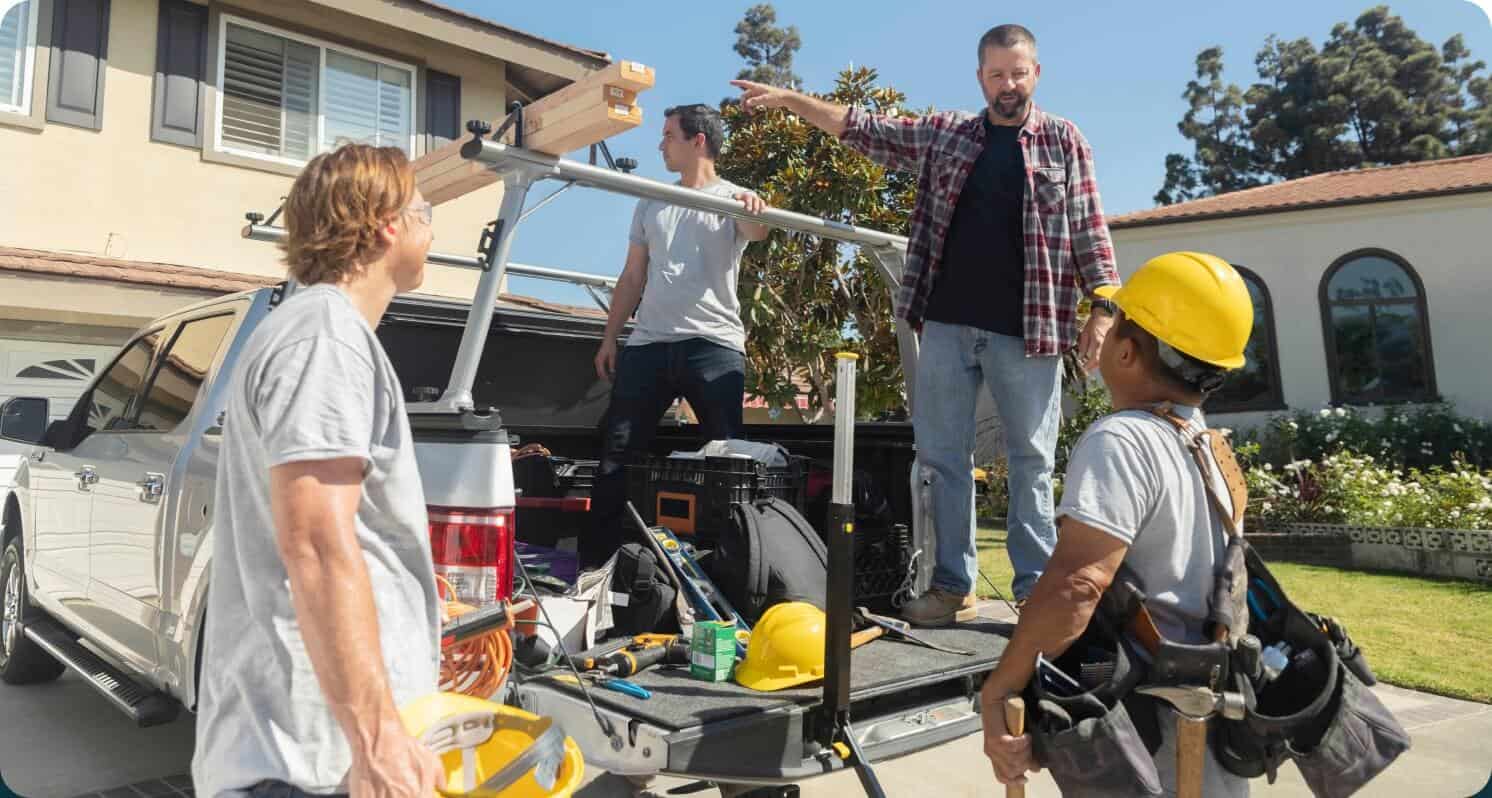 A team of 4 people gathering supplies out of a truck. Two of the people are standing in the truck and two men holding supplies on the ground. One person is pointing to something outside the frame.