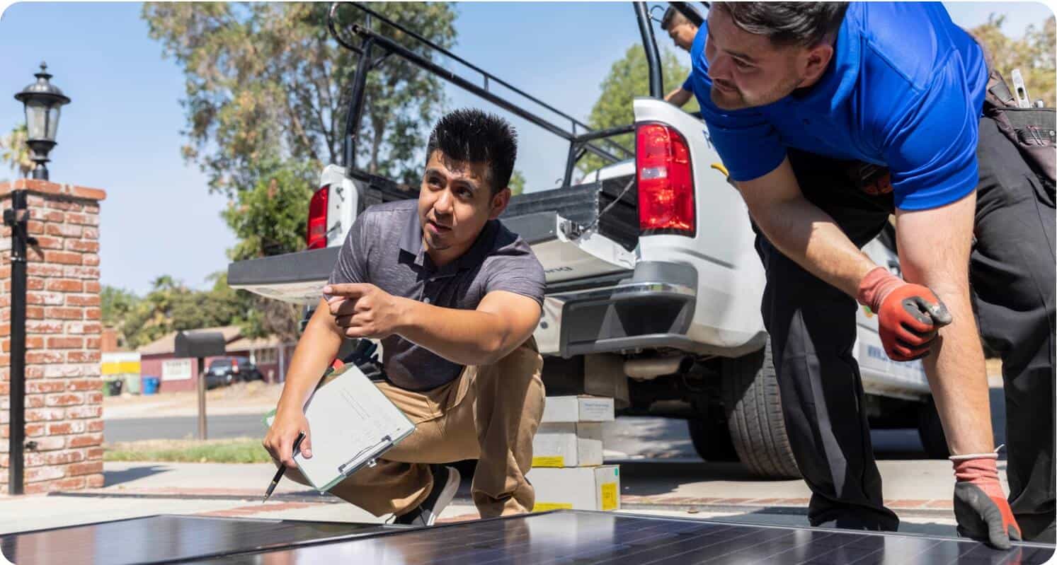 A team of three people working outside taking supplies off of a truck. One person is in the truck and the other two are on the ground. One man is lifting a large panel wile the other is pointing to the left and holding a clipboard.